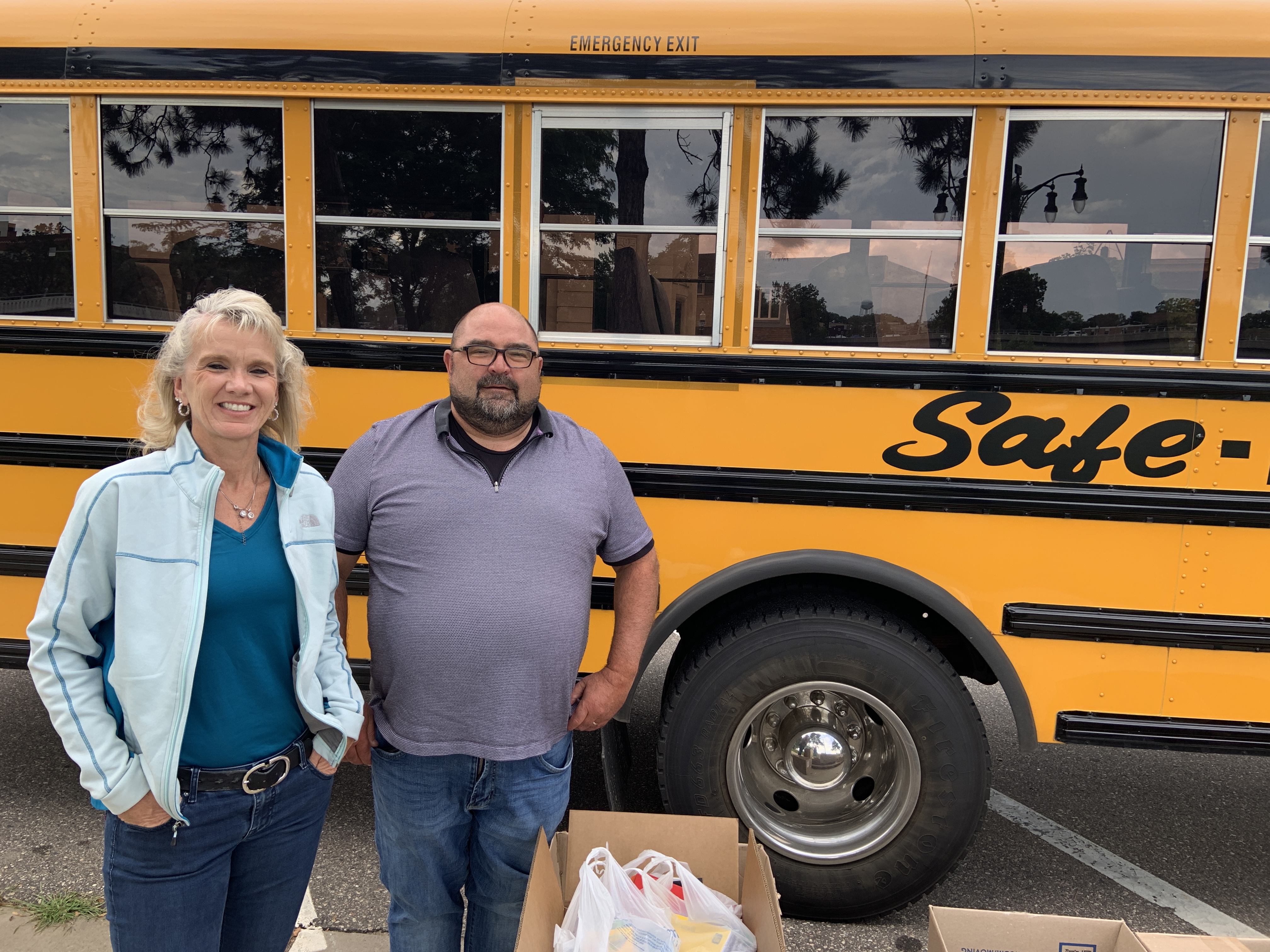 A man and a woman smiling in front of a school bus with some school supplies