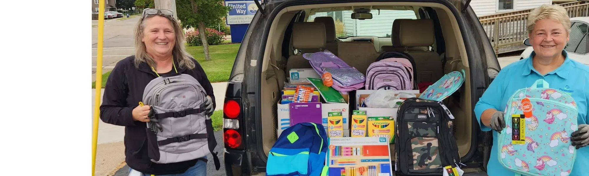 Two ladies holding backpacks in front of an open car trunk filled with school supplies.